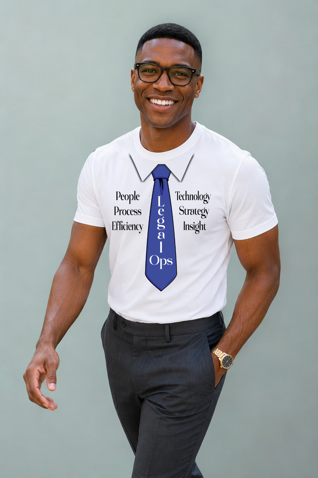 A smiling man wearing glasses, a white graphic T-shirt with a blue tie design and text, and dark trousers, standing against a plain light gray background.