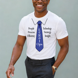 A smiling man wearing glasses, a white graphic T-shirt with a blue tie design and text, and dark trousers, standing against a plain light gray background.